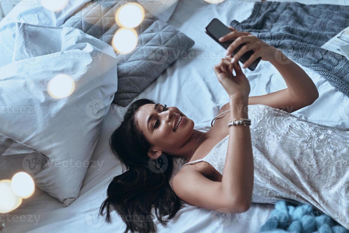conversando com o namorado. vista superior de uma jovem atraente usando telefone inteligente e sorrindo enquanto estava deitado na cama em casa foto