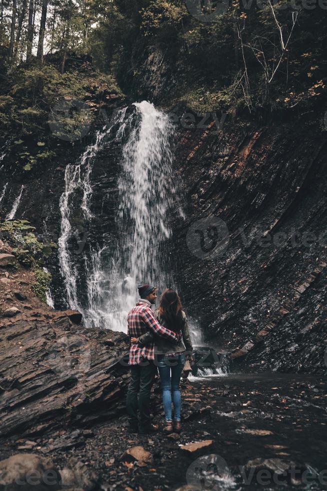 melhor viagem. vista traseira de corpo inteiro do jovem casal abraçando em pé perto da cachoeira foto