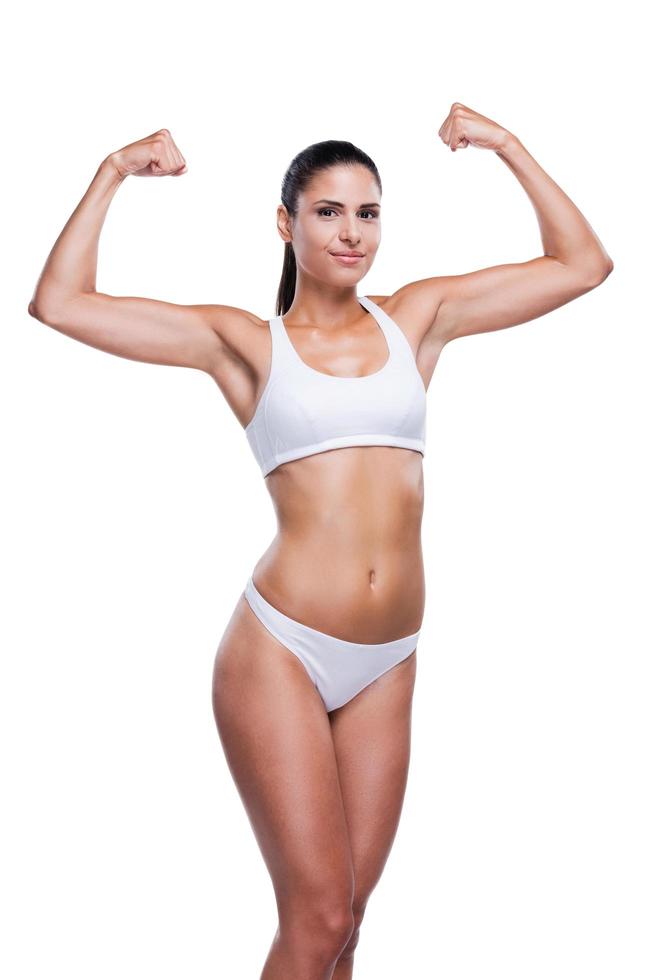 orgulhosa de seu corpo em forma. mulher jovem e bonita no sutiã branco e calcinha mostrando seu bíceps e sorrindo em pé isolado no branco foto