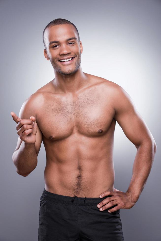 confiante em seu corpo perfeito. jovem africano sem camisa apontando para fora e sorrindo em pé contra um fundo cinza foto