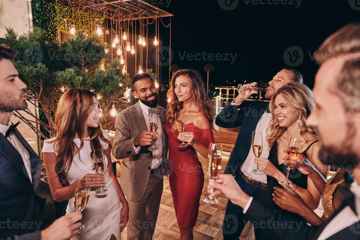 grupo de jovens bonitos em trajes formais se comunicando e sorrindo enquanto passa o tempo na festa de luxo foto