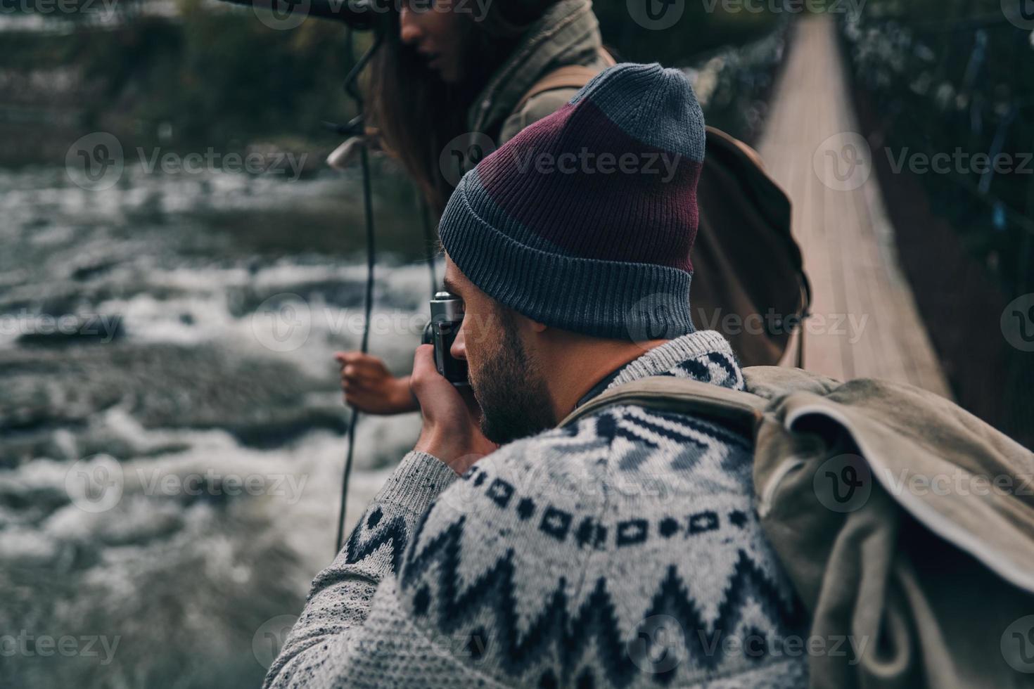 jovem moderno fotografando o rio em pé na ponte suspensa com sua namorada foto