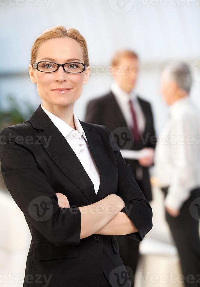 confiante em si mesma. alegre mulher madura em trajes formais, mantendo os braços cruzados e sorrindo enquanto seus colegas falando no fundo foto