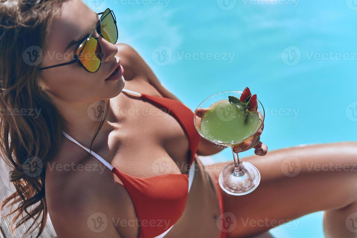 beleza sem esforço. vista superior de uma jovem atraente em trajes de banho segurando um copo enquanto está sentado à beira da piscina ao ar livre foto