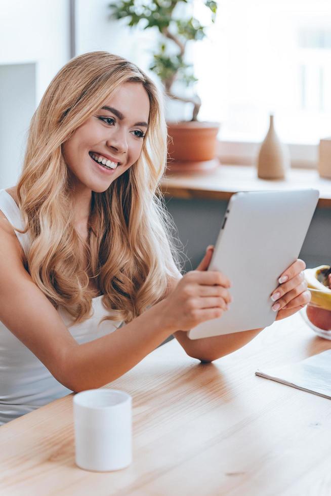 desfrutando de seu novo tablet digital. bela jovem alegre usando seu touchpad com sorriso enquanto está sentado na cozinha em casa foto