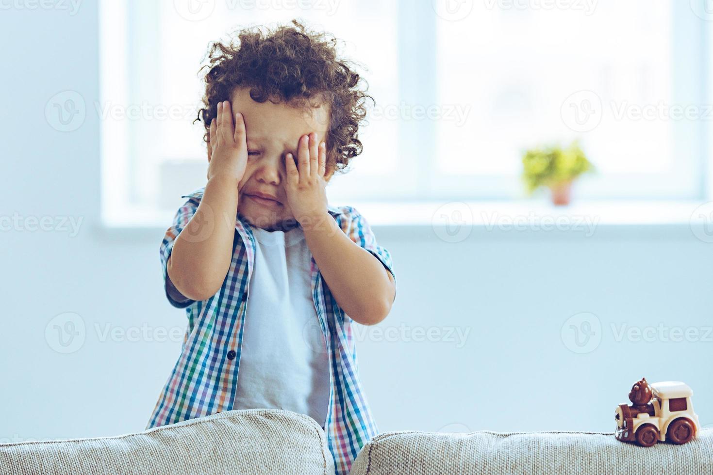 garotinho africano chorando e esfregando os olhos em pé no sofá em casa foto