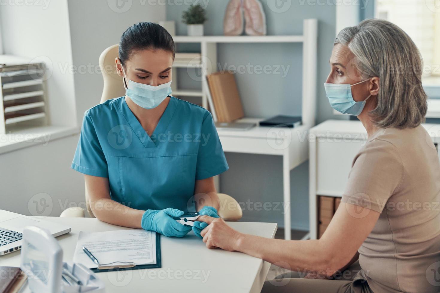 jovem enfermeira usando equipamento de monitoramento para verificar o pulso da mulher madura enquanto trabalhava no hospital foto