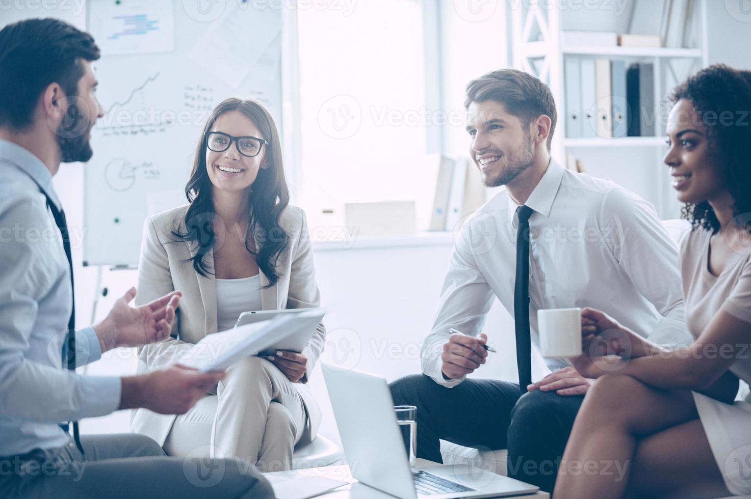 nosso trabalho é incrível jovem bonito segurando documentos e discutindo algo com seus colegas de trabalho com sorriso enquanto está sentado no sofá no escritório foto