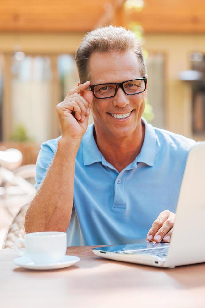 trabalhando com prazer. alegre homem maduro trabalhando no laptop e sorrindo enquanto está sentado à mesa ao ar livre com a casa ao fundo foto