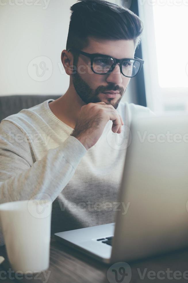 pensando em solução. jovem pensativo em óculos trabalhando no laptop e segurando a mão no queixo enquanto está sentado no escritório ou café foto