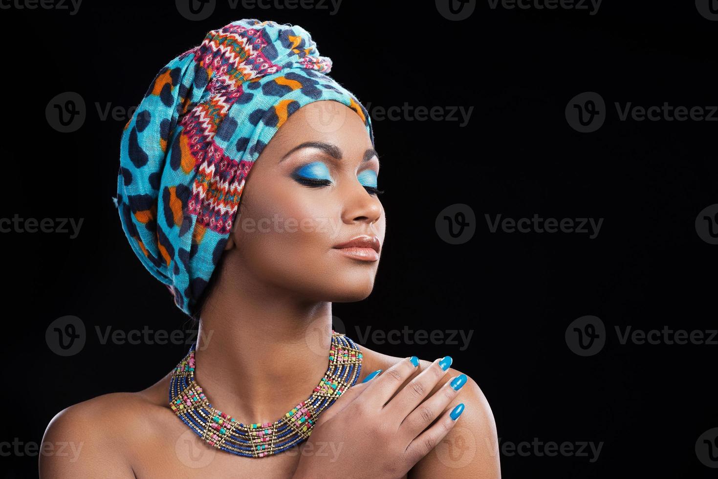 beleza africana. linda mulher africana vestindo um lenço na cabeça e colar mantendo os olhos fechados em pé contra um fundo preto foto