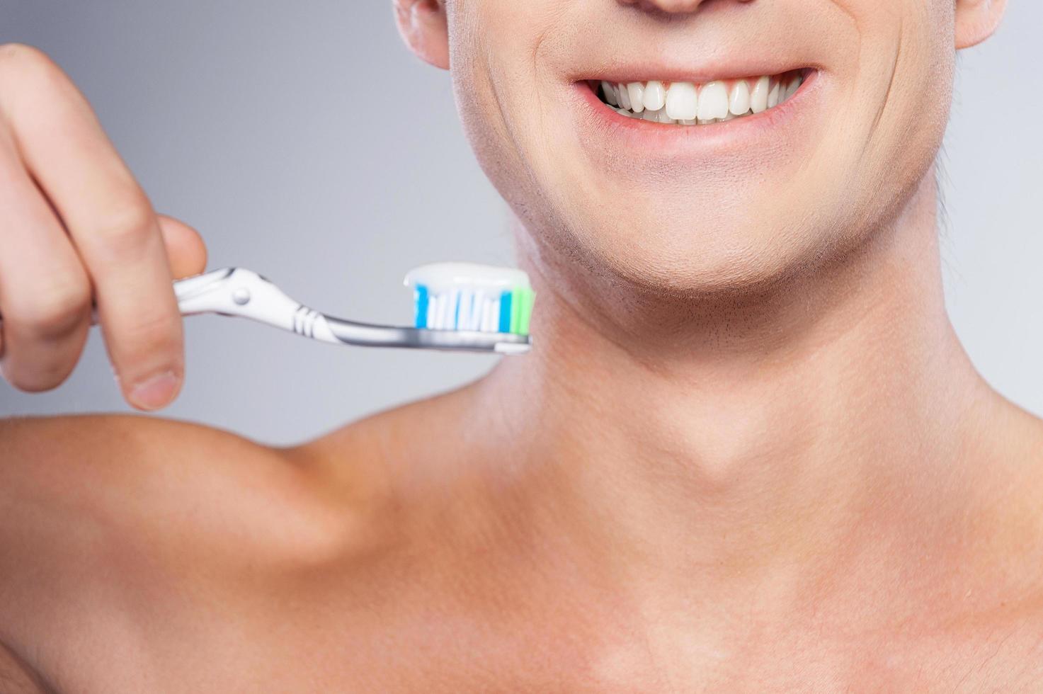 pronto para escovar os dentes. imagem recortada de jovem bonito sem camisa segurando uma escova de dentes com pasta de dente e sorrindo em pé contra um fundo cinza foto