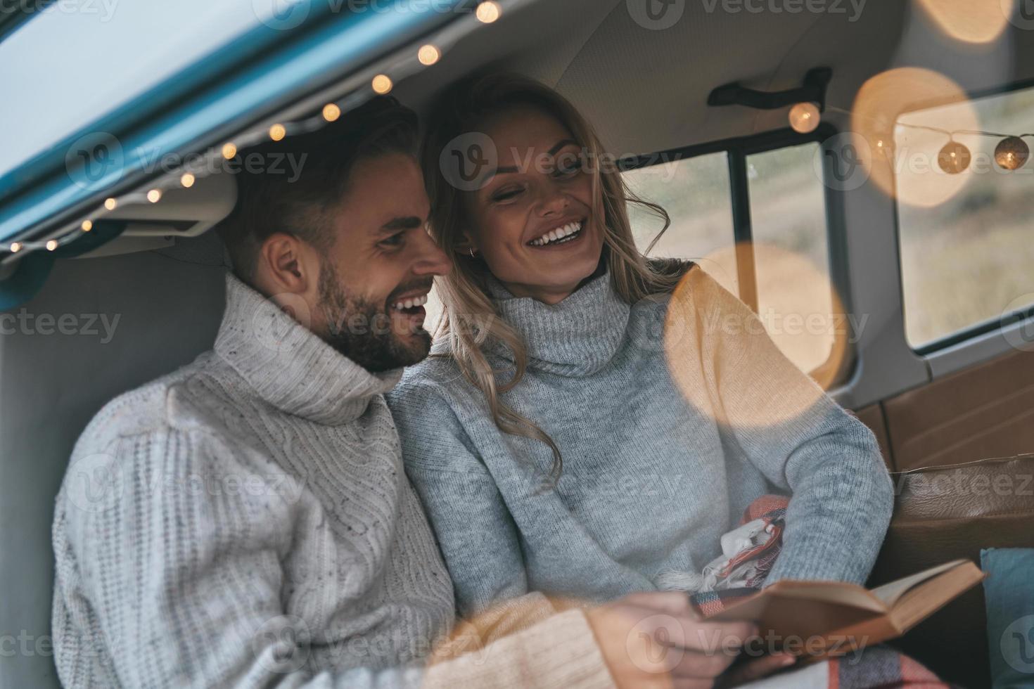 boa história. lindo casal jovem lendo o livro e sorrindo enquanto está sentado na mini van estilo retro foto