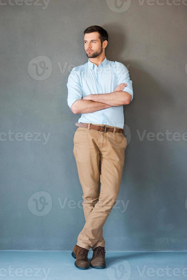 sério e confiante. comprimento total de jovem bonito na camisa, mantendo os braços cruzados e olhando para longe em pé contra um fundo cinza foto