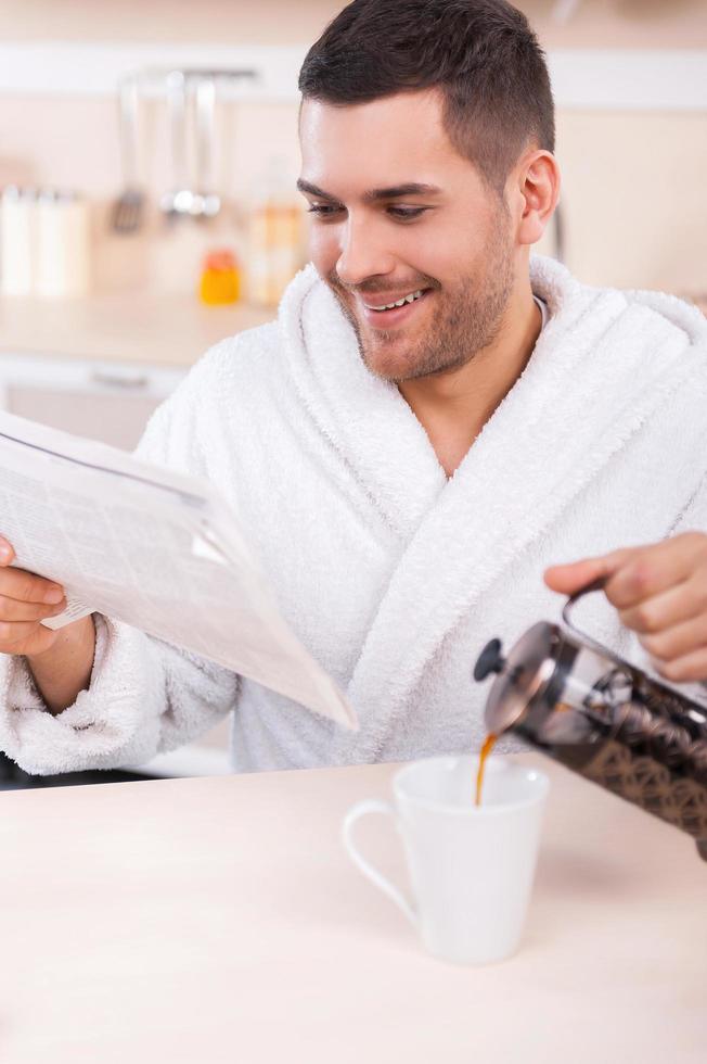 passando a manhã de domingo na cozinha. jovem bonito lendo jornal e servindo café na xícara foto