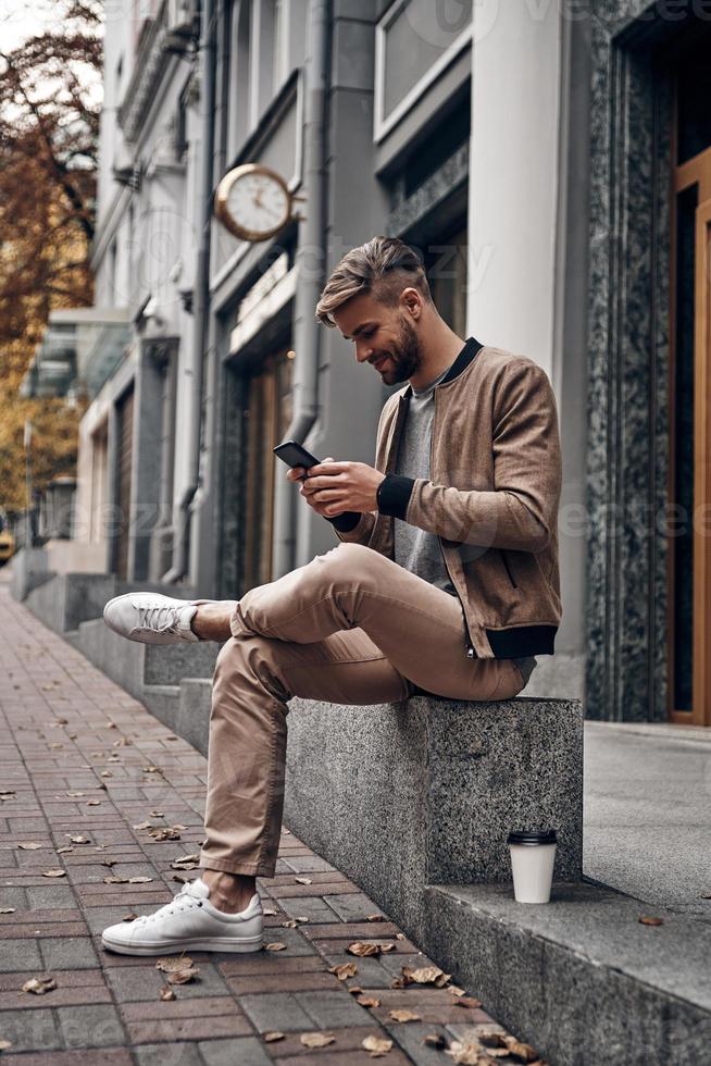 sempre online. jovem bonito em roupas casuais, usando seu telefone inteligente enquanto está sentado ao ar livre foto