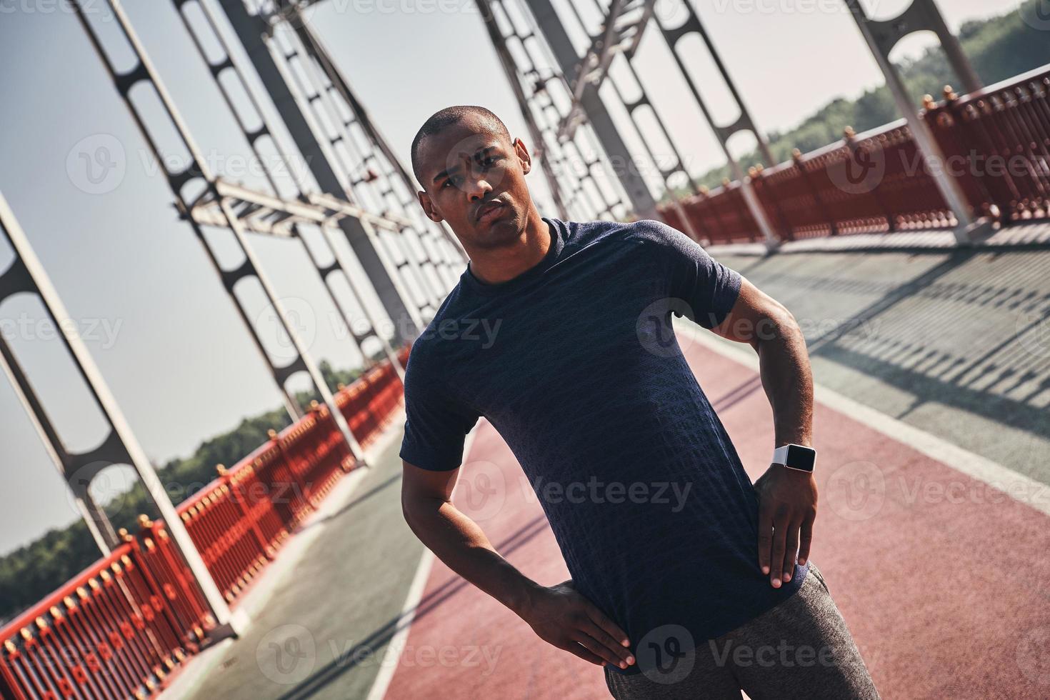 atleta forte. jovem africano bonito em roupas esportivas, olhando para a câmera em pé na ponte ao ar livre foto