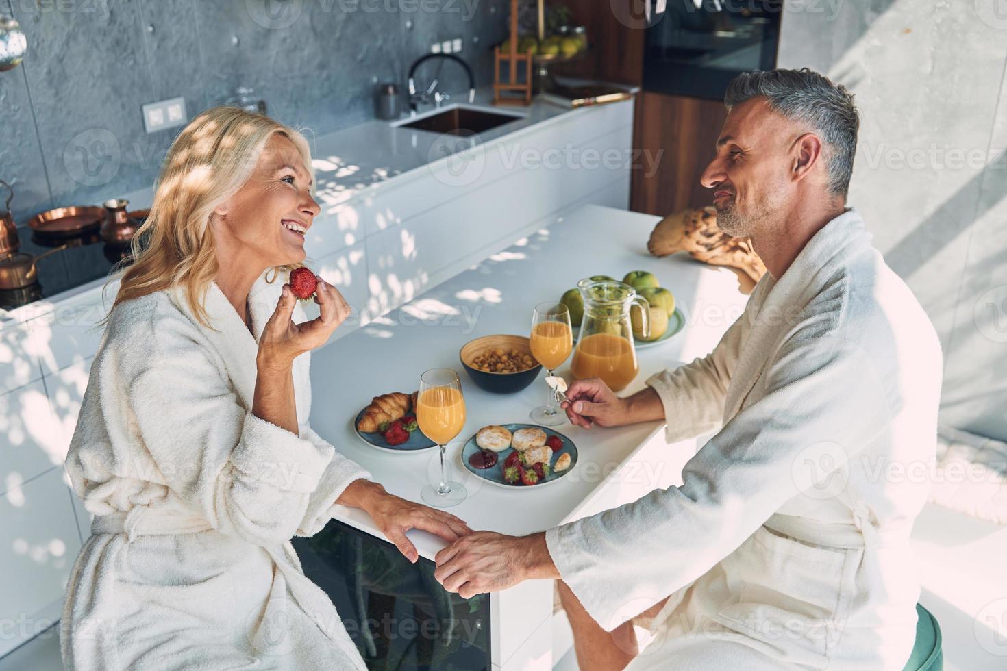 vista superior do lindo casal maduro em roupões de banho tomando café da manhã juntos enquanto passa o tempo na cozinha doméstica foto