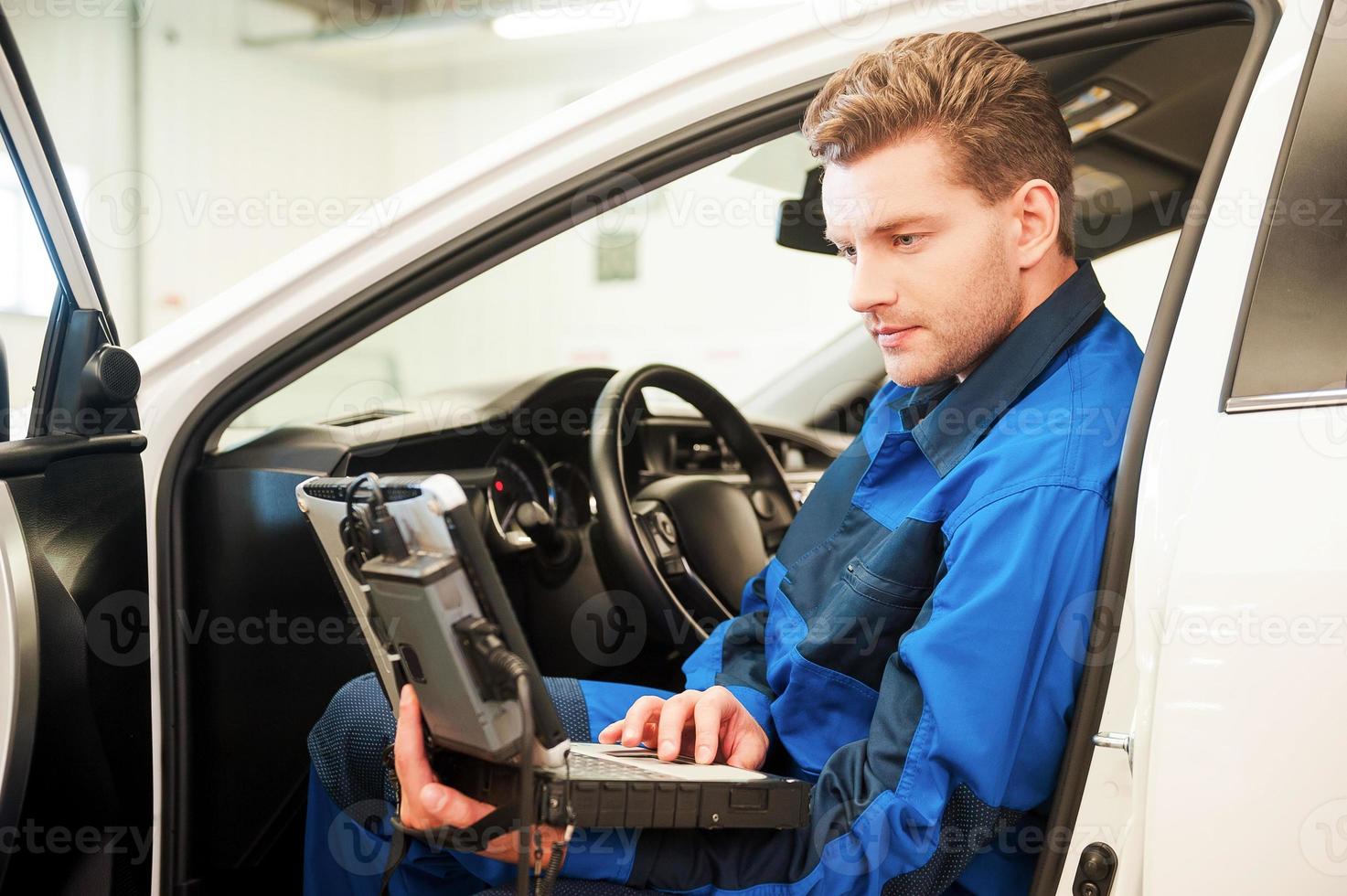 examinando o carro com computador especial. jovem confiante trabalhando no laptop especial enquanto está sentado em um carro na oficina foto