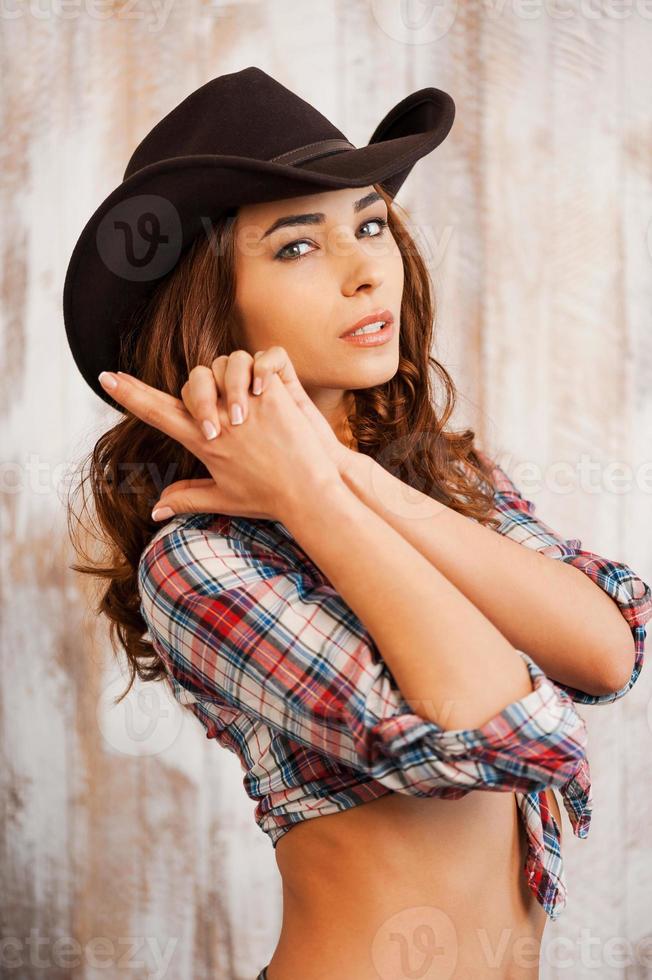 bonito e perigoso. bela jovem vaqueira gesticulando e olhando para a câmera em pé contra o fundo de madeira foto