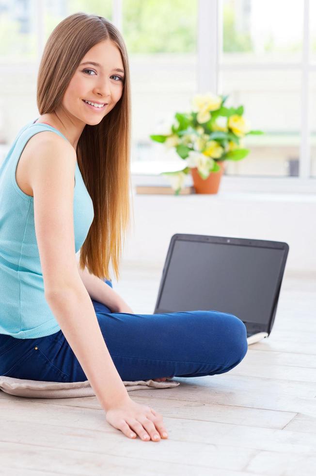 passar o tempo em casa. alegre adolescente usando computador e olhando por cima do ombro enquanto está sentado no chão em seu apartamento foto