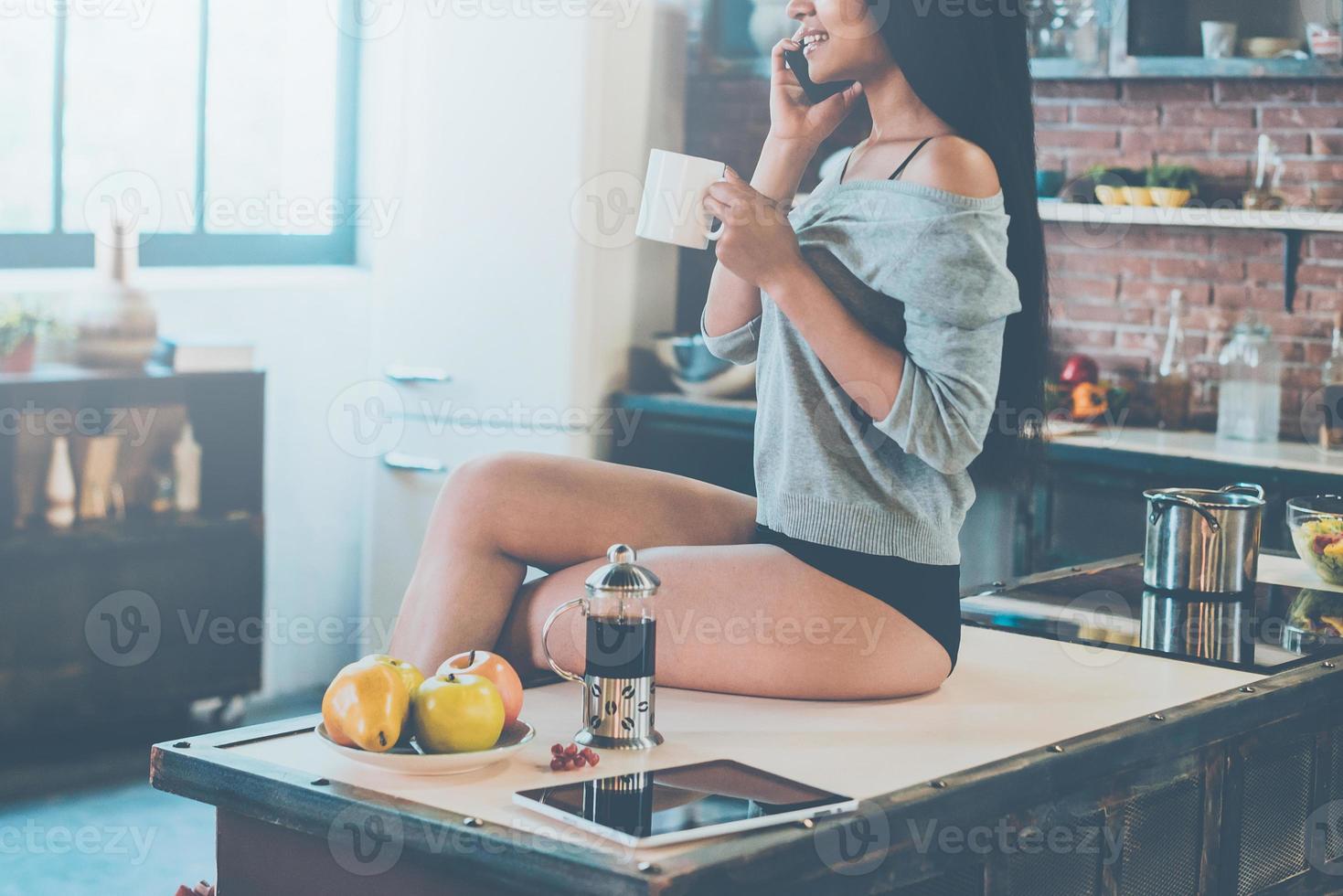 desfrutando de uma manhã despreocupada. close-up da bela jovem de raça mista, segurando a xícara de café e falando no celular com um sorriso enquanto está sentado na mesa da cozinha foto