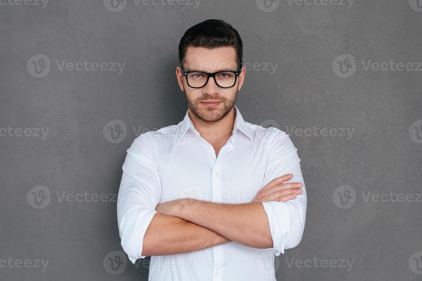 confiante e bem sucedido. jovem bonito abotoando sua camisa branca e olhando em pé contra um fundo cinza foto