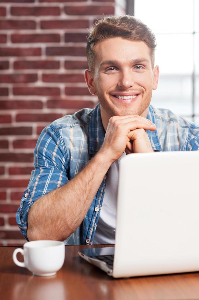 hora do café. jovem bonito segurando a xícara de café e sorrindo enquanto está sentado em seu local de trabalho foto