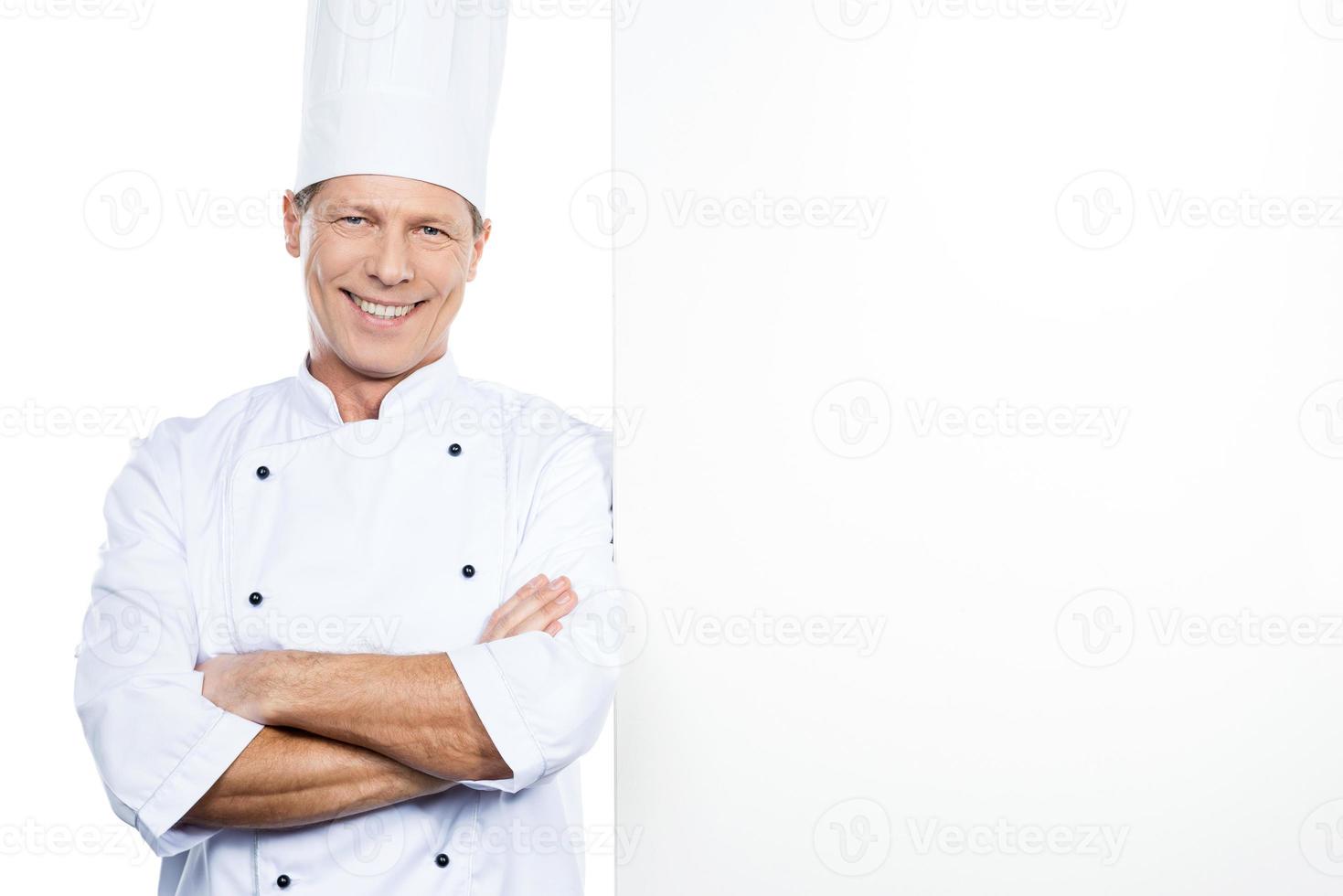 chef alegre. chef maduro confiante em uniforme branco, inclinando-se para o espaço da cópia e sorrindo em pé contra um fundo branco foto