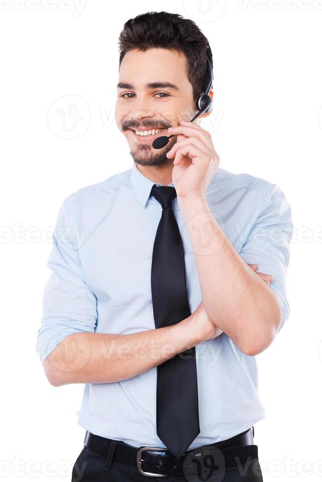 como posso ajudá-lo jovem e bonito operador masculino ajustando seu fone de ouvido e sorrindo em pé contra um fundo branco foto