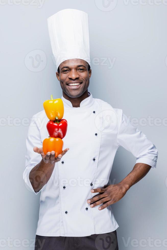 culinária colorida. confiante jovem chef africano em uniforme branco segurando pimentões multicoloridos e olhando para a câmera com sorriso em pé contra um fundo cinza foto