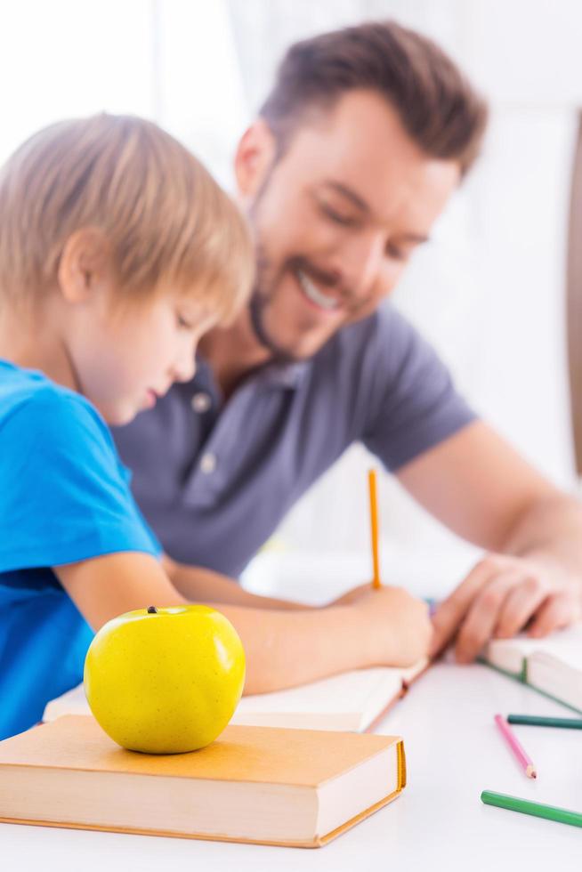 ajudando o filho com os trabalhos escolares. Vista lateral do jovem pai alegre ajudando seu filho com a lição de casa e sorrindo enquanto está sentado à mesa junto com a maçã verde em primeiro plano foto