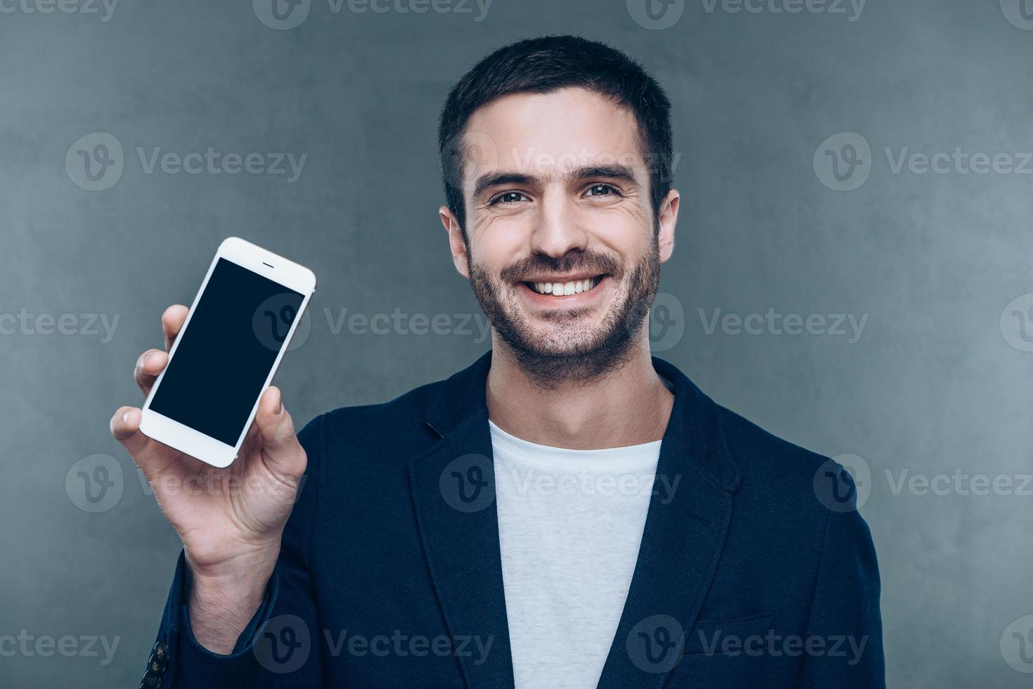 olhe para o meu novo telefone inteligente jovem alegre segurando o celular e sorrindo em pé contra um fundo cinza foto