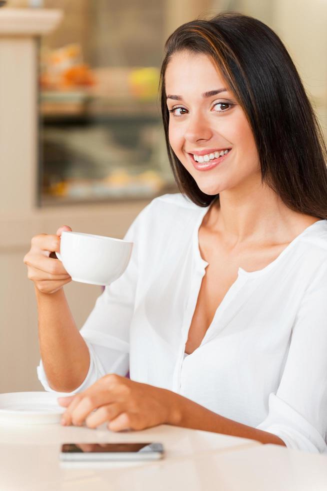 desfrutando de café fresco. bela jovem tomando café e sorrindo enquanto está sentado na cafeteria foto