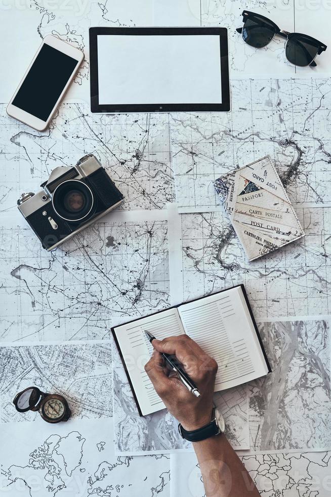 planejando viagens. close-up vista superior do homem escrevendo algo no diário com óculos de sol, câmera fotográfica, bússola, passaporte e telefone inteligente deitado no mapa ao redor foto