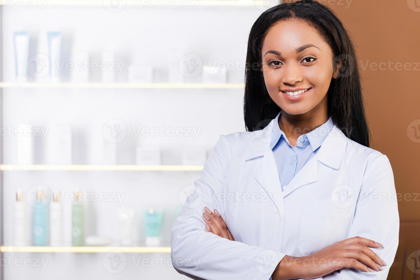 farmacêutico confiante. bela jovem africana em jaleco, mantendo os braços cruzados e sorrindo em pé na farmácia foto