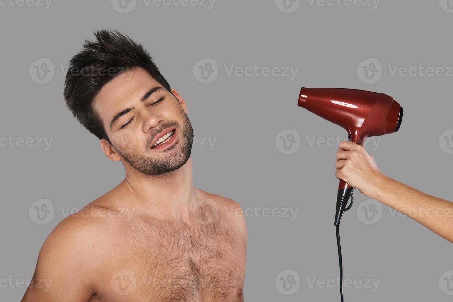 se sente bem. jovem bonito secando o cabelo e sorrindo em pé contra um fundo cinza foto