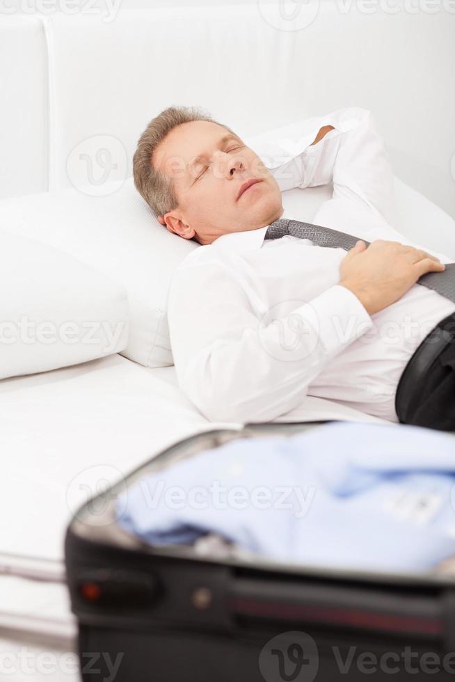 empresário cansado dormindo. homem de cabelo grisalho cansado de camisa e gravata deitado na cama e mantendo os olhos fechados enquanto a bagagem deitada na cama foto