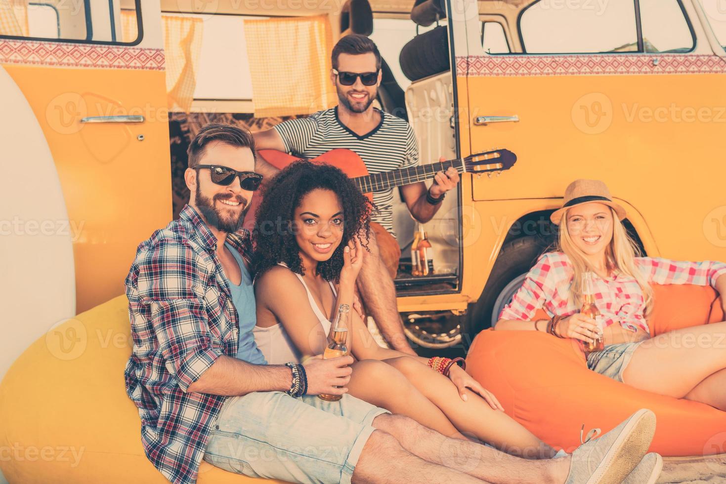 passar o verão juntos. três jovens felizes sentados perto de sua minivan com garrafas de cerveja enquanto homem tocando violão foto