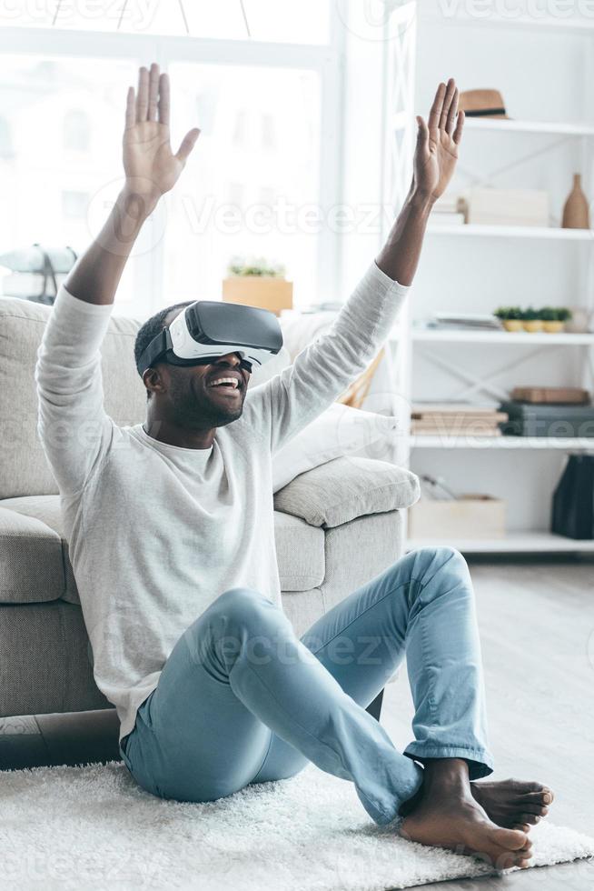 nova palavra de realidade virtual jovem africano bonito no fone de ouvido vr gesticulando e sorrindo enquanto está sentado no tapete em casa foto