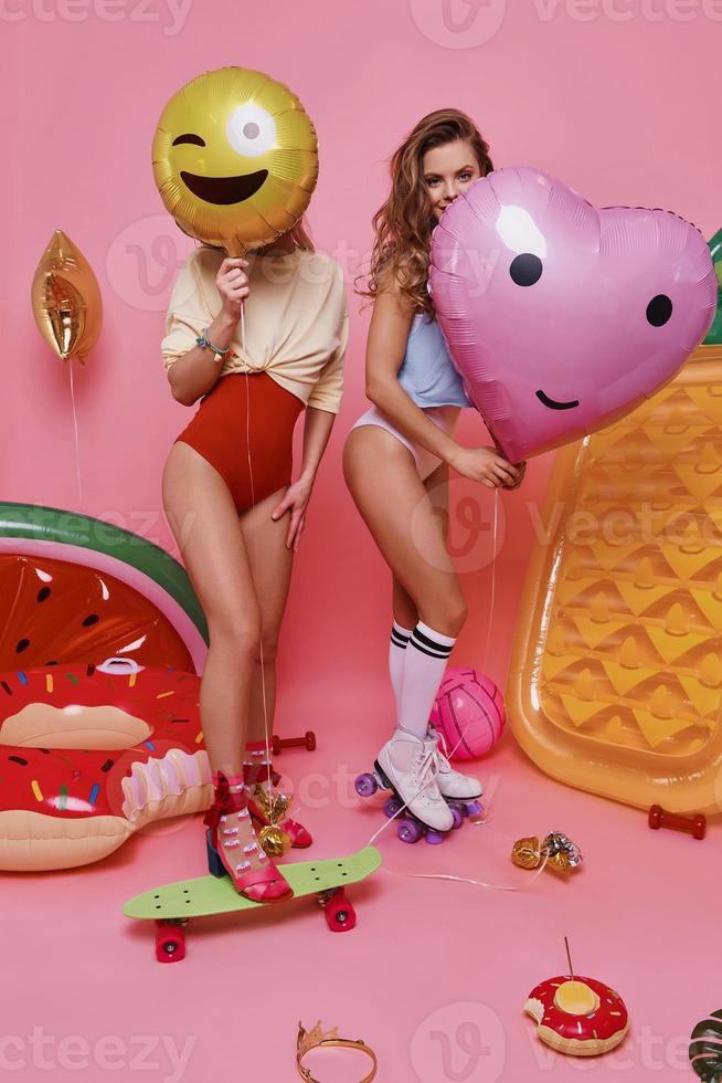 passar o tempo despreocupado. comprimento total de duas mulheres jovens em trajes de banho cobrindo rostos com balões em pé contra um fundo rosa foto