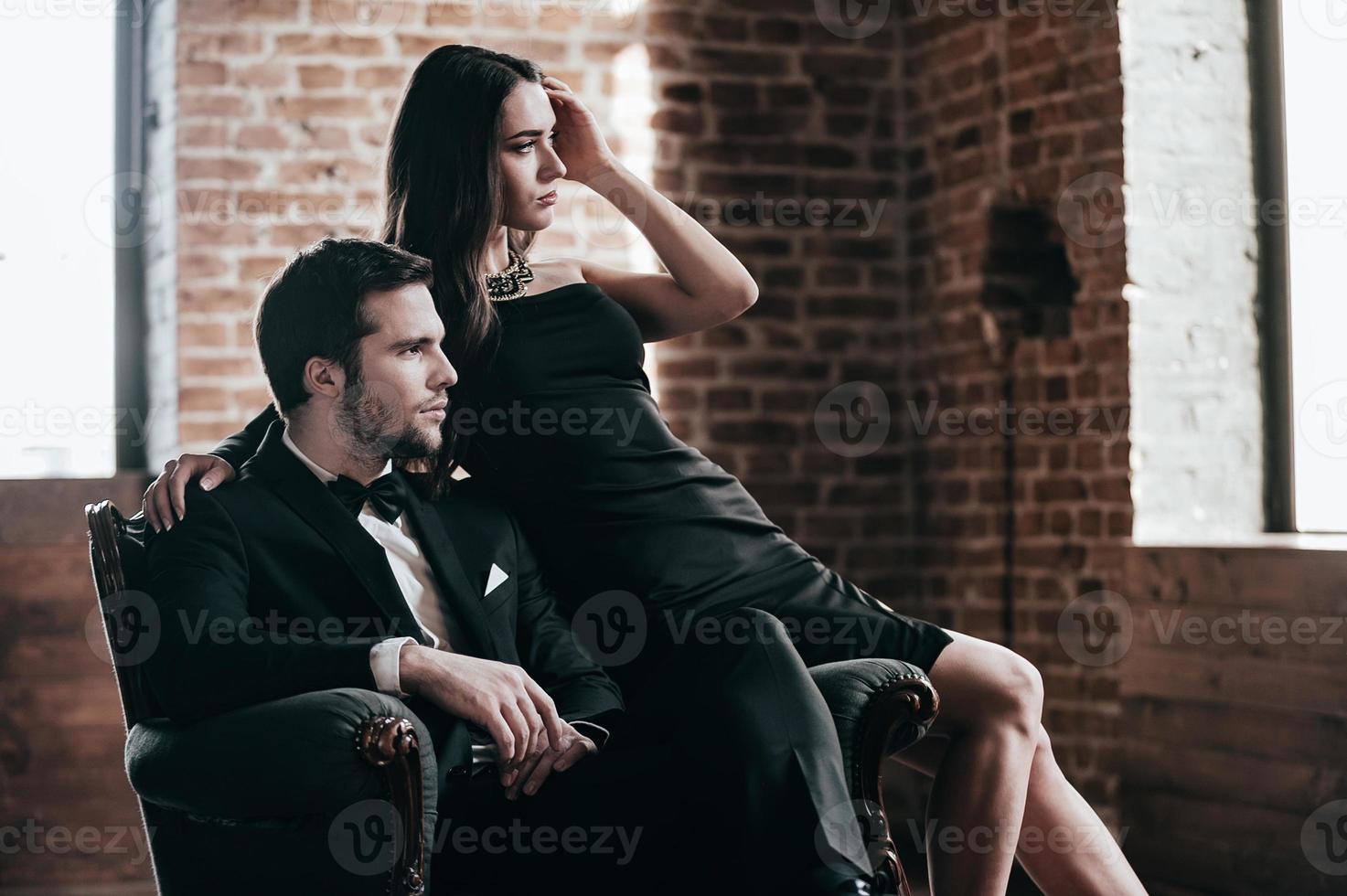 casal elegante. lindo casal jovem em trajes formais, olhando para a janela enquanto está sentado em uma cadeira juntos foto