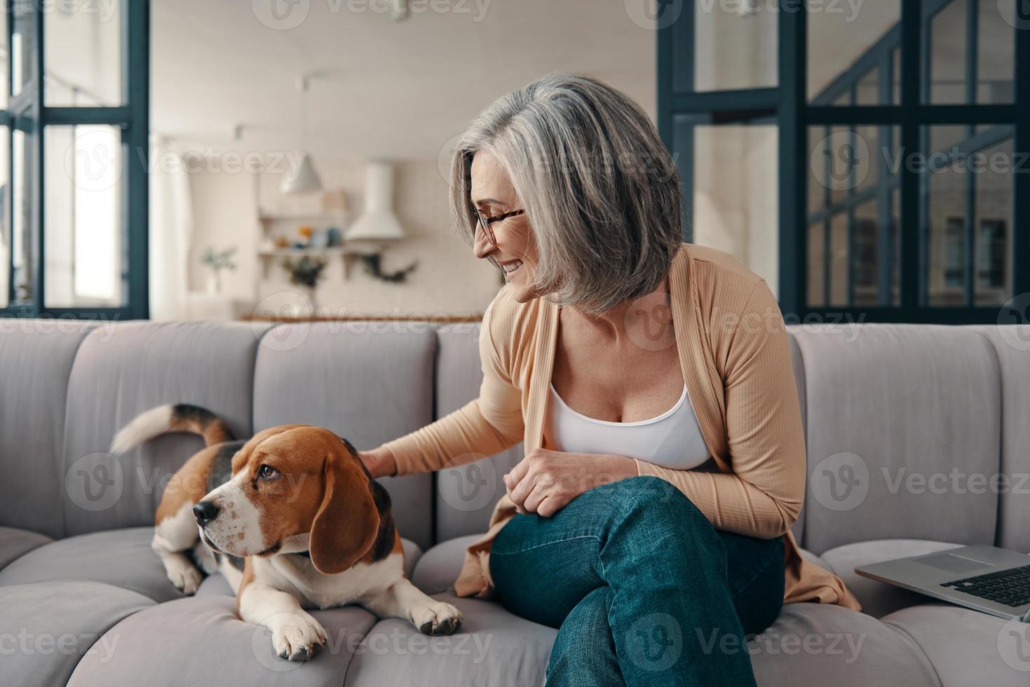 alegre mulher sênior em roupas casuais, passar tempo com seu cachorro enquanto está sentado no sofá em casa foto