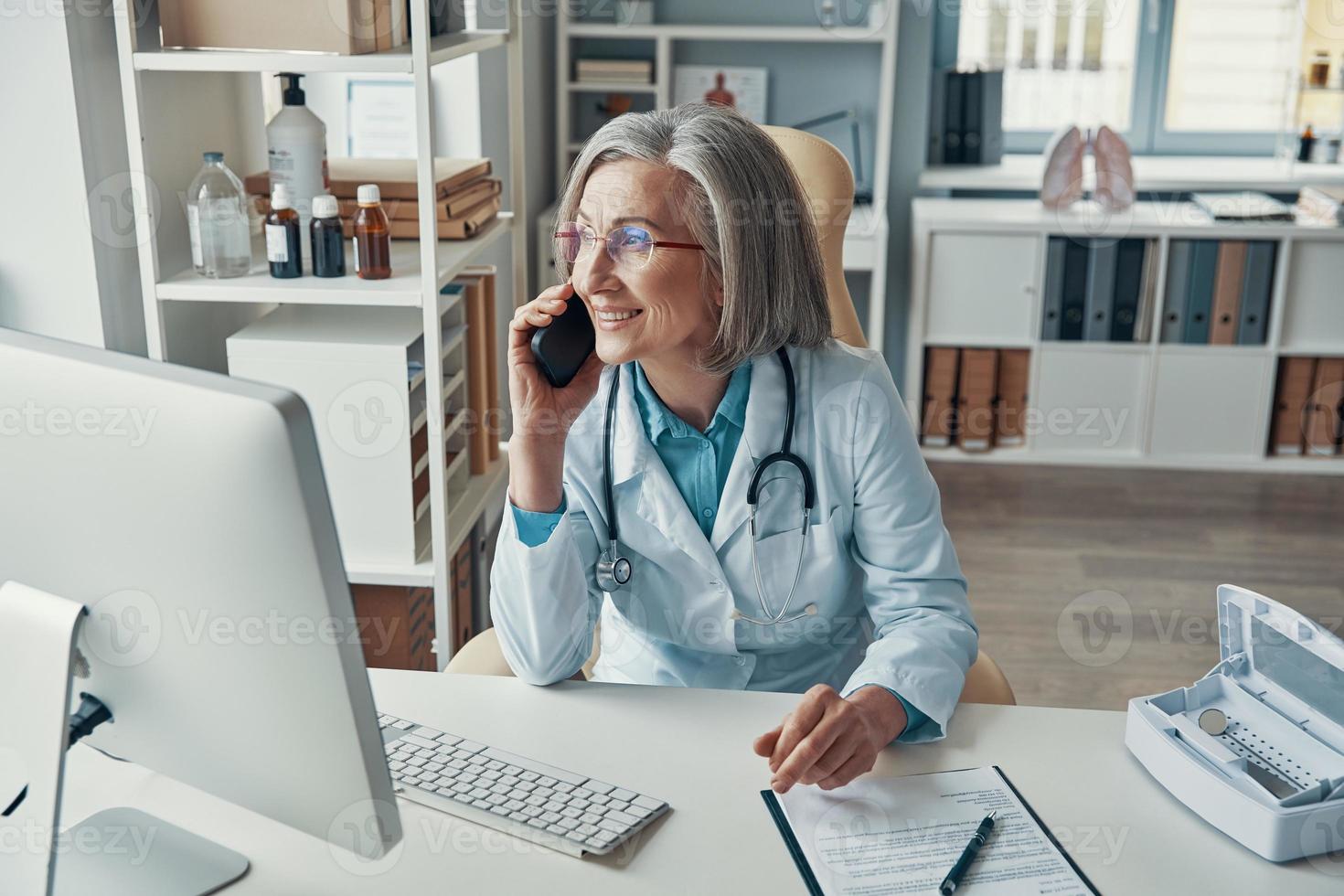 bela médica madura no jaleco branco falando ao telefone e sorrindo enquanto está sentado em seu escritório foto