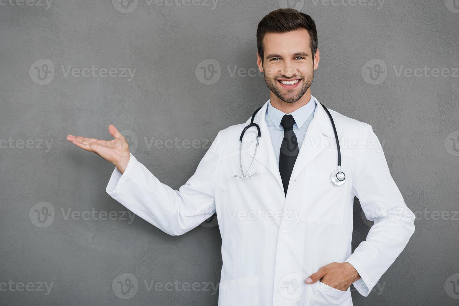 novas ideias para a medicina. alegre jovem médico de uniforme branco olhando para a câmera e apontando para fora em pé contra um fundo cinza foto