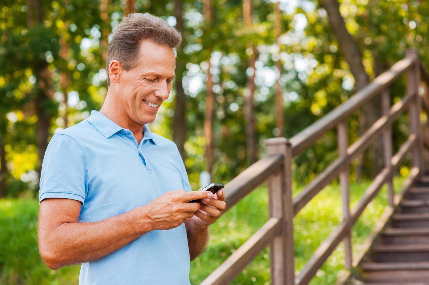examinando seu novo telefone inteligente. alegre homem maduro segurando o celular e olhando para ele com um sorriso em pé na escada de madeira foto