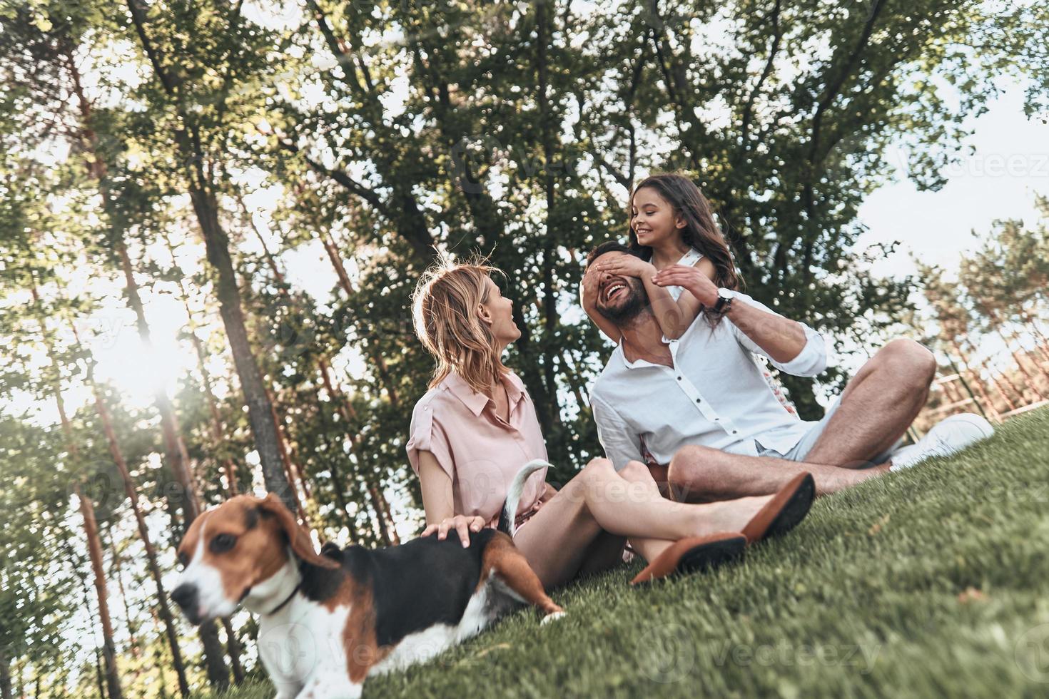momentos de alegria com a família. feliz jovem família de três com cachorro sorrindo enquanto está sentado na grama no parque foto