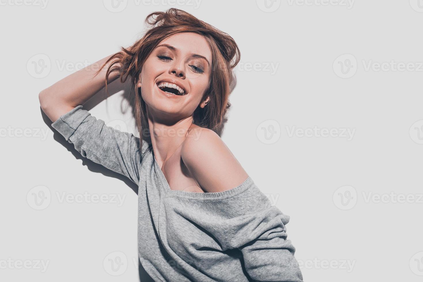 beleza feliz. mulher jovem e atraente sorridente segurando a mão no cabelo e mantendo os olhos fechados em pé contra um fundo cinza foto
