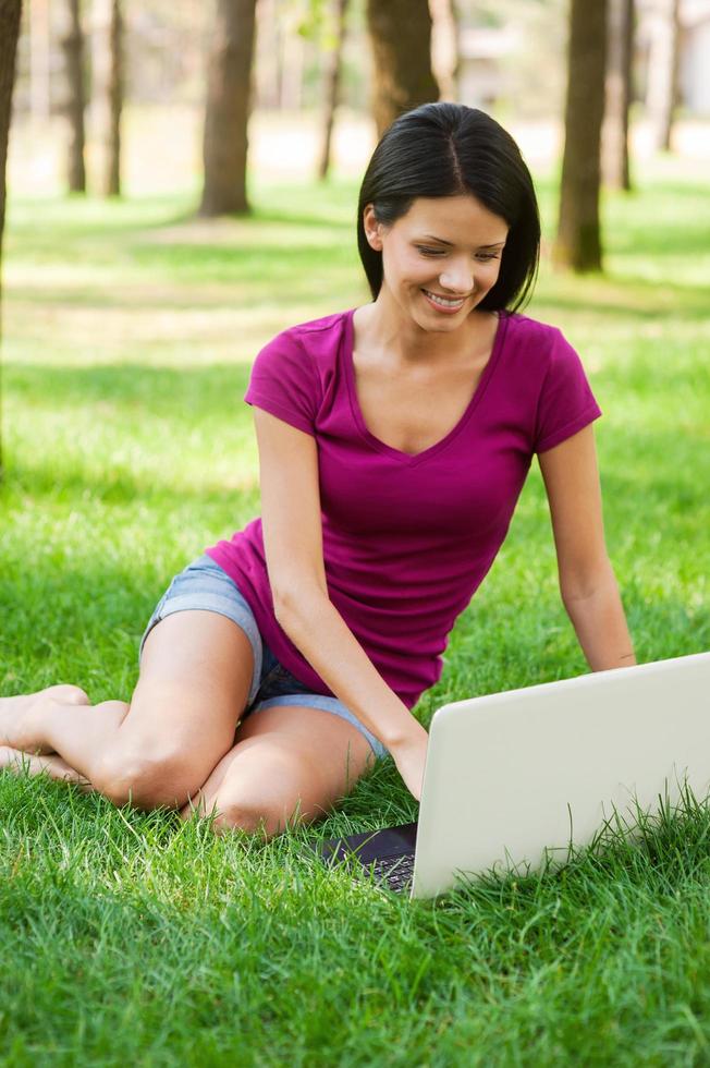 navegar na net ao ar livre. mulher jovem e atraente trabalhando no laptop e sorrindo enquanto estava deitado na grama foto