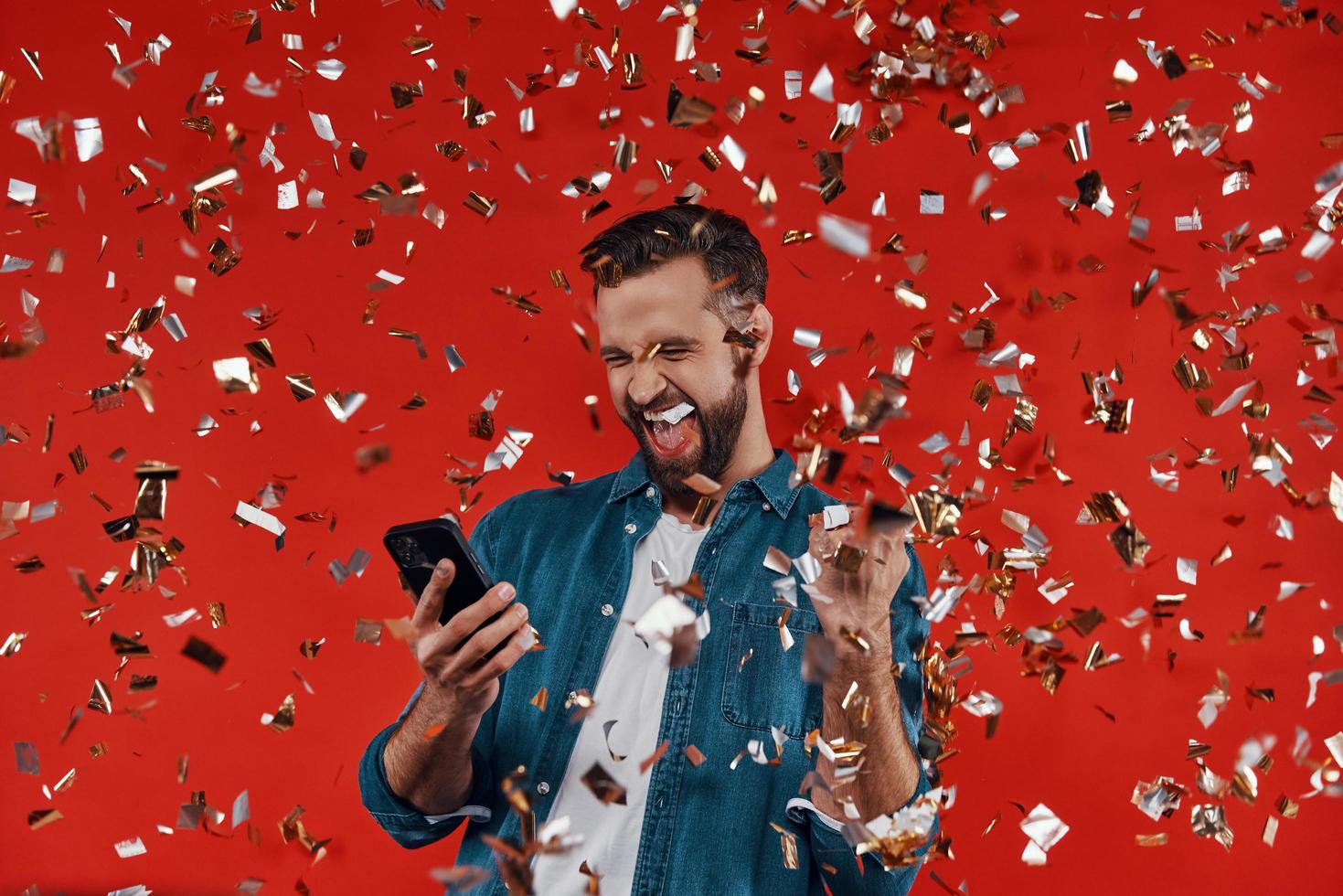 jovem feliz em roupas casuais, segurando o telefone inteligente e gesticulando em pé contra um fundo vermelho com confete voando ao redor foto